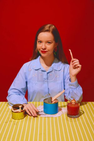 Foto de Joven hermosa mujer sentada a la mesa y comiendo carne enlatada, bebiendo jugo de tomate sobre fondo rojo. Representación de comida extraña. Concepto de arte pop fotografía, creatividad, comida, rareza - Imagen libre de derechos