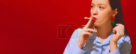 Téléchargez les photos : Jeune femme rousse aux lèvres rouges fumant partie poupée sur fond rouge. Fumer pour le plaisir, arrêter la mauvaise habitude. Concept de photographie pop art, créativité, surréalisme, bizarrerie - en image libre de droit