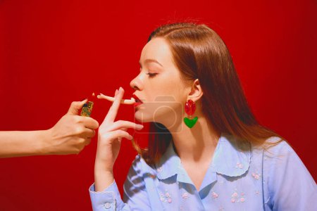 Foto de Mujer pelirroja joven con labios rojos parte de muñeca humeante sobre fondo rojo. Fumar por diversión, dejar el mal hábito. Concepto de arte pop fotografía, creatividad, surrealismo, rareza - Imagen libre de derechos