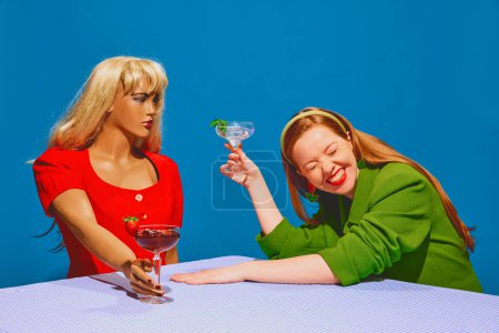 Photo for Young redhead positive woman smiling, drinking cocktail and talking to mannequin against blue background. Communication with inner self. Concept of pop art photography, creativity - Royalty Free Image