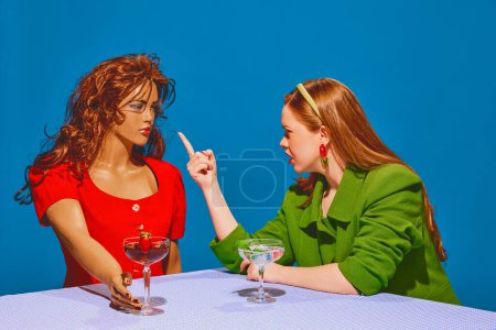 Young redhead woman drinking cocktail, talking, arguing with mannequin against blue background. Intense communication. Concept of pop art photography, creativity, surrealism, psychology