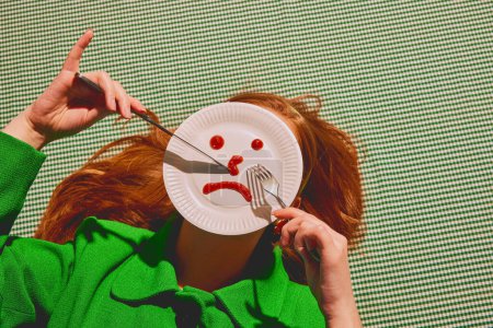 Photo for Young redhead woman lying with knife and fork over paper plates showing sad face. Choosing mood for today. Concept of pop art photography, creativity, psychology, surrealism - Royalty Free Image