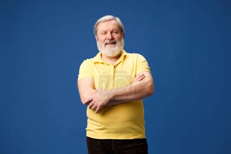 Foto de Retrato de anciano barbudo sonriente en polo amarillo de pie con artes cruzadas contra fondo azul del estudio. Salud y bienestar. Concepto de emociones humanas, estilo de vida, moda casual - Imagen libre de derechos