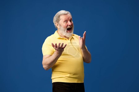 Foto de Anciano barbudo vestido de polo amarillo, expresando emoción positiva contra el fondo azul del estudio. Ventas, noticias, sorpresa. Concepto de emociones humanas, estilo de vida, moda casual - Imagen libre de derechos