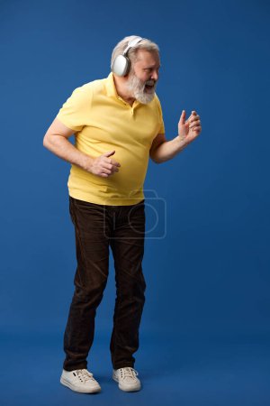 Foto de Anciano barbudo en polo amarillo escuchando música en auriculares y bailando sobre fondo azul del estudio. De larga duración. Concepto de emociones humanas, estilo de vida, moda casual - Imagen libre de derechos
