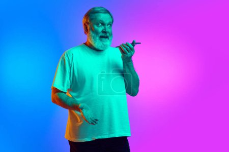 Photo for Emotional bearded senior man recording voice message via mobile phone on gradient blue-purple background in neon light. Concept of human emotions, lifestyle, casual fashion - Royalty Free Image