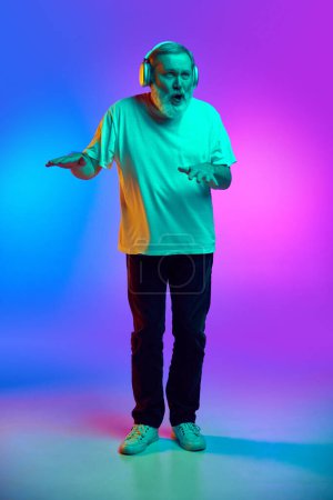 Téléchargez les photos : Homme âgé barbu en vêtements décontractés écoutant de la musique dans les écouteurs et dansant sur fond dégradé bleu-violet sous la lumière du néon. Concept d'émotions humaines, style de vie, mode décontractée - en image libre de droit