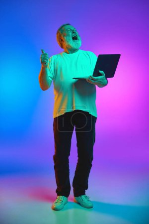 Foto de Longitud completa del hombre mayor de pie con el ordenador portátil con la expresión de la felicidad y el éxito en el fondo azul-púrpura degradado en luz de neón. Concepto de emociones humanas, estilo de vida, moda casual - Imagen libre de derechos