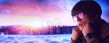 Junger Mann mit Brille, mit nachdenklichem Gesichtsausdruck auf dem winterlichen Sonnenuntergang, verschneite Landschaft. Optische Markenwerbung, die stylische Brillen für jede Jahreszeit hervorhebt.