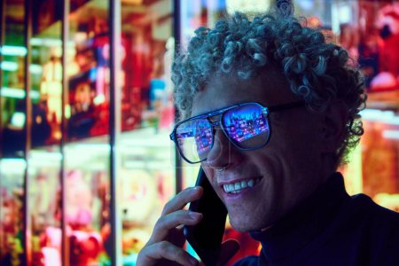 Téléchargez les photos : Homme bouclé souriant debout près d'un grand centre commercial de la ville dans des lunettes avec réflexion des lumières urbaines dans des lunettes et parler sur un téléphone mobile. Scène de ville du soir. Concept de style de vie urbain, technologie - en image libre de droit