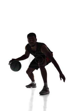 Foto de Silueta de joven concentrado, jugador de baloncesto en movimiento durante el juego, bola de goteo aislado sobre fondo blanco. Concepto de deporte profesional, competición, juego, torneo, acción - Imagen libre de derechos