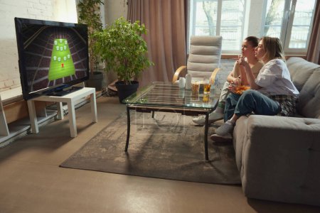 Photo for Tense game moment. Two girls, friends sitting in living room at home, eating snack and watching online football match on TV. Concept of sport, championship, leisure and entertainment - Royalty Free Image