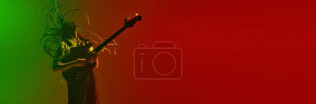 Foto de Hombre emocional, músico con rastas tocando la guitarra eléctrica contra el degradado fondo verde rojo en luz de neón. Concepto de música, performance, festival, concierto. Banner, anuncio - Imagen libre de derechos
