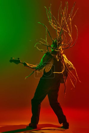 Photo for Young energetic musician with long dreadlocks playing electric guitar with passion and energy against gradient red green background in neon light. Concept of music, performance, festival, concert - Royalty Free Image