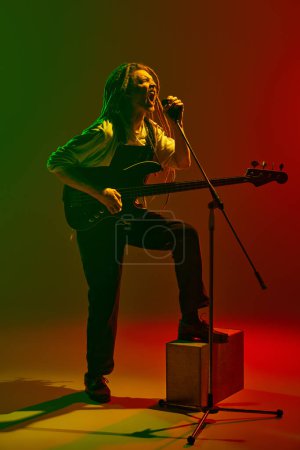 Photo for Expressive musician with dreadlocks playing electric guitar and singing on microphone on stage against gradient red green background in neon light. Concept of music, performance, festival, concert - Royalty Free Image