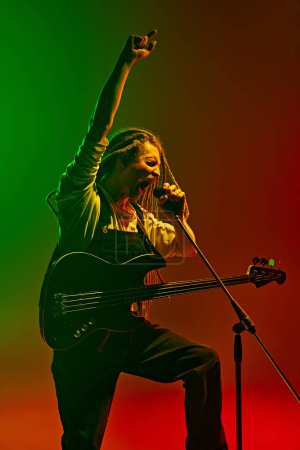 Photo for Energetic soulful man with dreadlocks, musician with electric guitar singing against gradient red green background in neon light. Concept of music, performance, festival, concert - Royalty Free Image