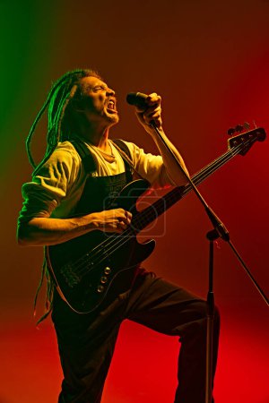 Photo for Young man, expressive singer with dreadlocks emotionally singing, performing solo against gradient red green background in neon light. Concept of music, performance, festival, concert - Royalty Free Image