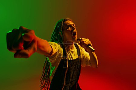 Photo for Connecting with audience. Young artistic and talented man with dreadlocks signing in microphone against gradient red green background in neon light. Concept of music, performance, festival, concert - Royalty Free Image
