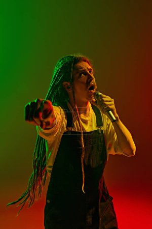 Photo for Emotional atmosphere created by man with dreadlocks showing unforgettable vocal performance against gradient red green background in neon light. Concept of music, performance, festival, concert - Royalty Free Image