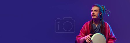 Foto de Música country. Joven músico artístico, hombre con rastas tocando tambores bongo sobre fondo púrpura en luz de neón. Concepto de música, performance, festival, concierto. Banner, anuncio - Imagen libre de derechos