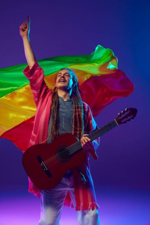 Foto de Músico enérgico, hombre con llamativos rastas tocando la guitarra sobre la bandera roja, amarilla y verde sobre fondo púrpura en luz de neón. Concepto de música, performance, festival, concierto - Imagen libre de derechos