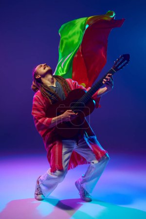 Foto de Artista dinámico, hombre emocional con rastas creando música, tocando la guitarra sobre fondo púrpura en luz de neón con bandera roja, amarilla, verde. Concepto de música, performance, festival, concierto - Imagen libre de derechos