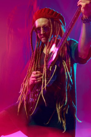 Photo for Charismatic young man with dreadlocks, in sunglasses and beanie passionately strumming an electric guitar against pink background in neon light. Concept of music, performance, festival, concert - Royalty Free Image
