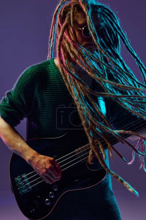 Foto de Retrato de tipo elegante con rastas, músico tocando la guitarra sobre fondo púrpura oscuro en luz de neón. Perdido en la música. Concepto de música, performance, festival, concierto - Imagen libre de derechos