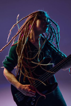 Photo for Passionate performer with dreadlocks pouring his heart out through his guitar playing against dark purple background in neon light. Concept of music, performance, festival, concert - Royalty Free Image