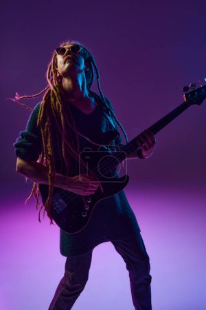 Foto de Artista apasionado con rastas derramando su corazón a través de su guitarra tocando sobre fondo púrpura oscuro en luz de neón. Concepto de música, performance, festival, concierto - Imagen libre de derechos