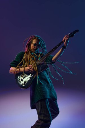 Photo for Soulful musician, charismatic guitarist with dreadlocks playing guitar, performing solo against dark purple background in neon light. Concept of music, performance, festival, concert - Royalty Free Image