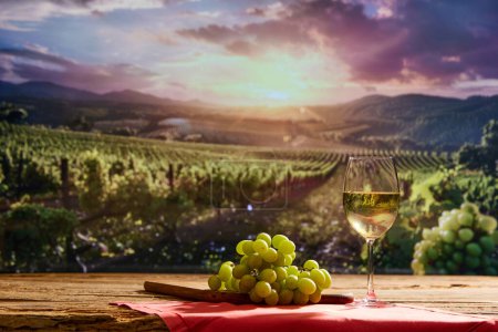 Glass of white wine and bunch of grapes on a wooden table. Blurred vineyard with rows of grapevine on background. Concept of winemaking, organic beverage, nature, traditions