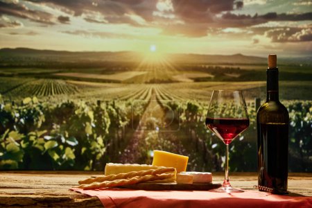 Photo for Glass of red wine and bottle on wooden table with cheese and breadsticks with vineyard during sunset time on background. Concept of winemaking, organic beverage, nature, traditions - Royalty Free Image