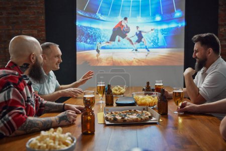 Foto de Amigos sentados juntos, inmersos en la emoción de un partido de fútbol en línea transmitido por la televisión, con cerveza y aperitivos en la mesa. Discusión del juego. Concepto de deporte, campeonato, aficionados al deporte, ocio - Imagen libre de derechos