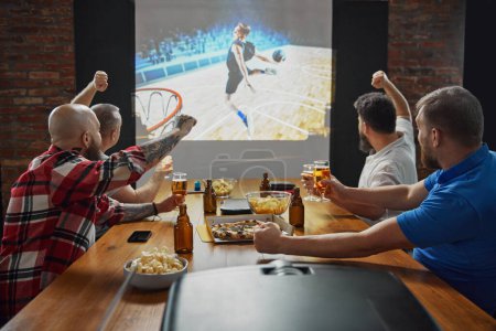 Photo for Men, friends sitting at table with beer and snacks, watching online basketball game translation on TV and cheering up favorite team. Concept of sport, championship, game, sport fans, leisure - Royalty Free Image