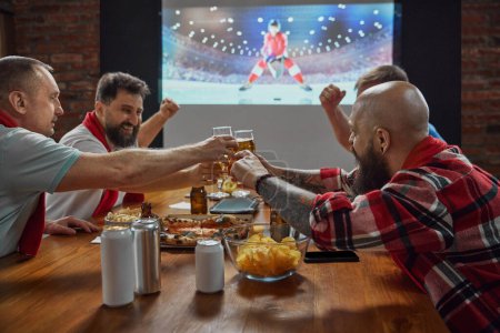Foto de Los entusiastas de los deportes, amigos que se reúnen en casa para ver el partido de hockey en línea en la televisión, con cerveza y aperitivos amplificando la emoción. Concepto de deporte, campeonato, juego, aficionados al deporte, ocio - Imagen libre de derechos