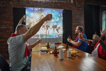 Foto de Amigos se reúnen para ver un partido de fútbol americano en línea en la televisión, con cerveza y aperitivos, expresando el disfrute del juego. Concepto de deporte, campeonato, juego, aficionados al deporte, ocio - Imagen libre de derechos