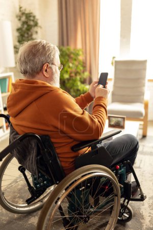 Foto de Anciano sentado en silla de ruedas en la sala de estar en casa y utilizando el teléfono móvil para la comunicación, el ocio y la lectura de noticias. Concepto de salud, estilo de vida, bienestar, comodidad, rehabilitación - Imagen libre de derechos