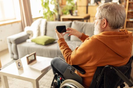 Téléchargez les photos : Homme âgé assis en fauteuil roulant à la maison dans le salon et regarder des nouvelles, des vidéos sur téléphone mobile. Utilisation de gadget pour les besoins quotidiens. Concept de santé, mode de vie, bien-être, confort, autonomisation - en image libre de droit