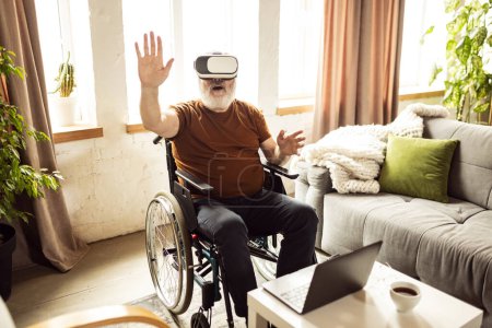 Téléchargez les photos : Explorer de nouveaux gadgets numériques. Homme âgé assis en fauteuil roulant, portant des lunettes VR, prenant des aventures virtuelles à la maison. Concept de santé, mode de vie, bien-être, confort, autonomisation - en image libre de droit