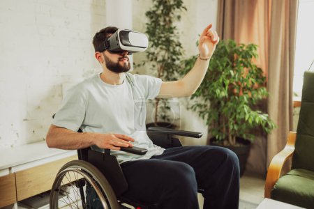 Foto de Joven sentado en silla de ruedas en la sala de estar en casa, con gafas VR y explorando las noticias de tecnología innovadora. Concepto de salud, estilo de vida, bienestar, comodidad, empoderamiento - Imagen libre de derechos