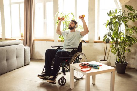 Photo for Young man sitting in wheelchair in headphones, training, doing exercises with dumbbells at home. Staying active at home. Concept of healthcare, lifestyle, wellness, comfort, empowerment - Royalty Free Image