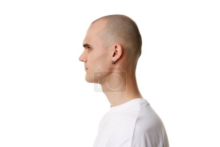 Téléchargez les photos : Côté, vue de profil du jeune homme à tête chauve et visage rasé, percement d'oreille debout en t-shirt blanc isolé sur fond blanc. Concept de soins de la peau, beauté masculine, bien-être, cosmétique, auto-soin - en image libre de droit