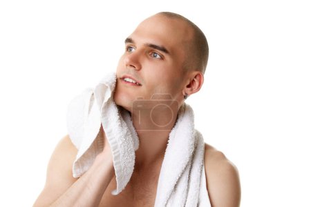 Téléchargez les photos : Jeune homme chauve avec serviette après avoir lavé le visage isolé sur fond de studio blanc. La routine du matin. Concept de soins de la peau, beauté masculine, bien-être, cosmétique, auto-soin - en image libre de droit