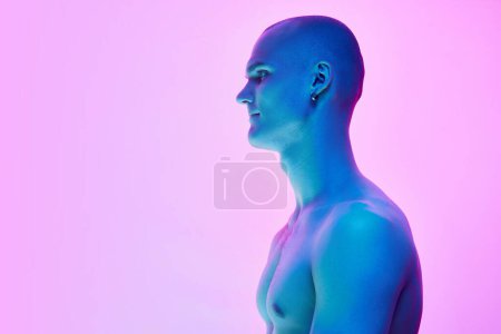 Téléchargez les photos : Image vue de côté du jeune homme chauve au corps musclé torse nu posant sur fond rose bleu dégradé sous la lumière du néon. Concept de beauté masculine, corps, jeunesse, fitness, sport, santé - en image libre de droit