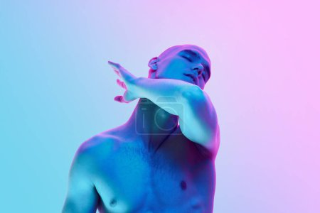 Téléchargez les photos : Portrait de jeune homme chauve torse nu posant avec des gestes sur fond rose bleu dégradé au néon. Concept de beauté masculine, corps, jeunesse, fitness, sport, santé - en image libre de droit