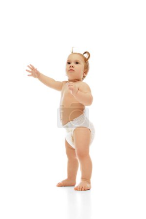 Photo for Curious little baby girl, toddler, kid in diaper, with two ponytails standing with raised arm isolated on white background. Exploring world. Concept of childhood, care, health, well-being, parenthood - Royalty Free Image