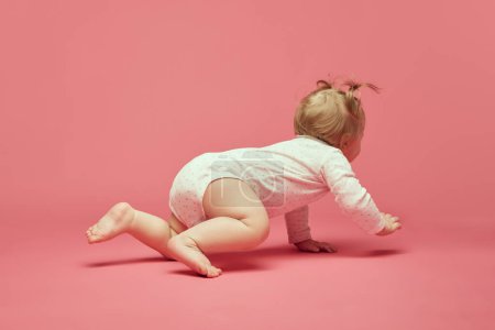 Téléchargez les photos : Souriant petite fille heureuse, tout-petit dans un confortable onesie rampant, jouant sur fond rose. Joyeux temps. Concept d'enfance, soins, santé, bien-être, parentalité - en image libre de droit