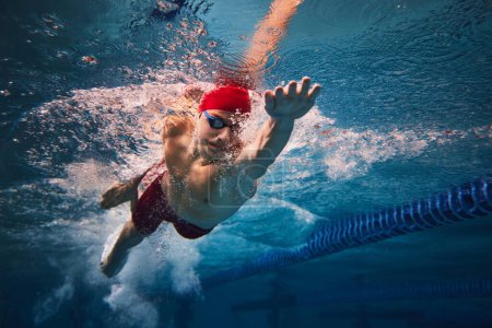 Photo for Resilience and strength. Concentrated young man in red cap and goggles in motion, swimming in pool, training. Concept of professional sport, health, endurance, strength, active lifestyle - Royalty Free Image