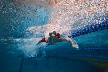 Photo for Strength and resilience. Young man, swimmer in red cap and goggles swimming, training in pool indoors. Concept of professional sport, health, endurance, competition, active lifestyle - Royalty Free Image